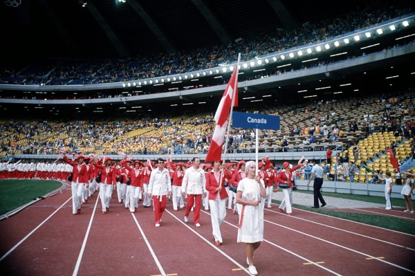 Team Canada in red and white outfits marching behind flag and sign