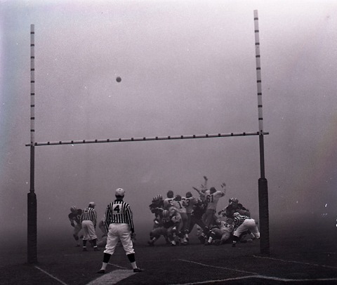 photograph of football above goalposts with players and referee in fog